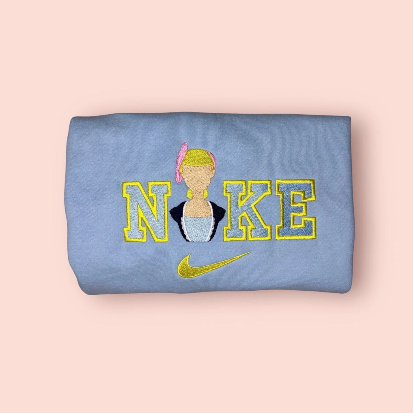 Sudadera de color azul celeste pastel con un delicado bordado de Bo Peep (Betty la Pastorcita) de Toy Story. Diseño estilo universitario "Nike bootleg" con letras amarillas y la silueta de la heroína de porcelana. Esta prenda personalizada de edición coleccionista es el regalo ideal para fans de Disney y Pixar. Tejido suave y confortable, perfecta para outfits femeninos y dulces.