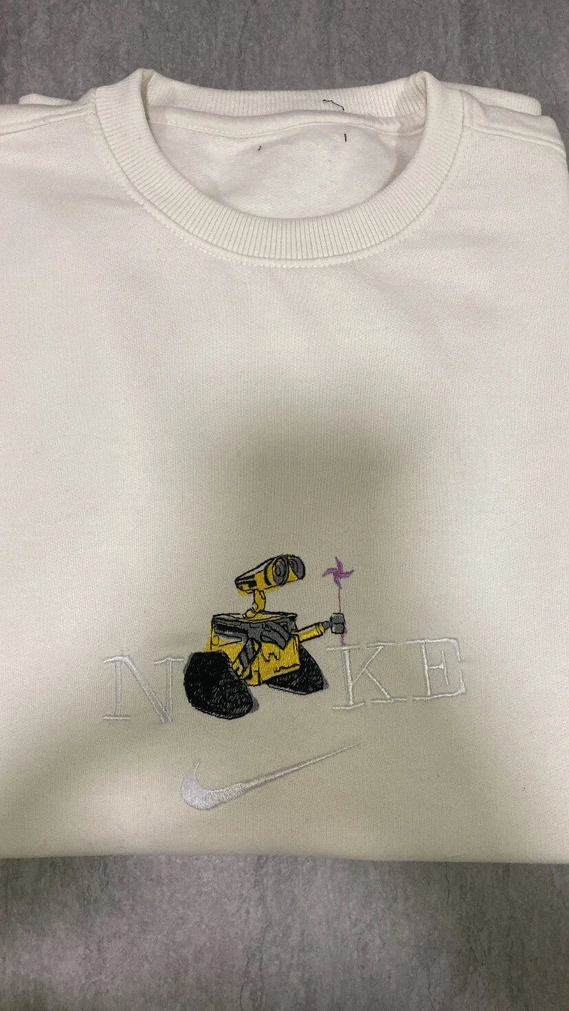Sudadera de color crema o hueso con un bordado detallado del robot WALL-E. El diseño muestra al tierno personaje de Pixar con sus ojos curiosos y sosteniendo un molinillo de viento, integrado en un logo de estilo deportivo. Ropa personalizada de alta calidad, perfecta para fans de Disney Pixar. La pieza esencial para completar el dúo con la sudadera de Eva (EVE) y crear el 'matching outfit' más romántico de la galaxia.