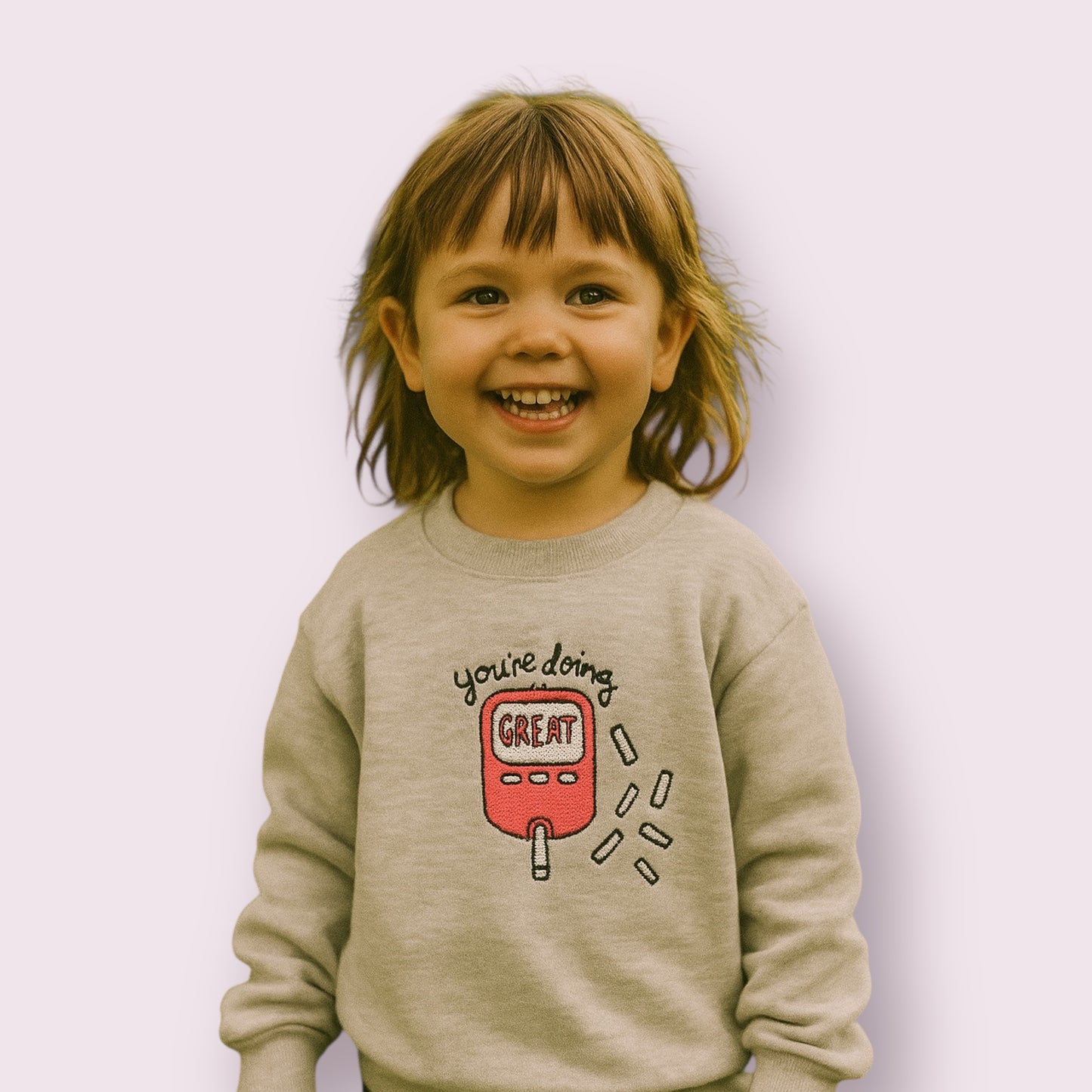 Niña sonriente con sudadera infantil gris claro con diseño de glucómetro rosa y mensaje motivador "You're doing great", ropa para niños con diabetes tipo 1, ideal para visibilizar la diabetes con estilo y ternura