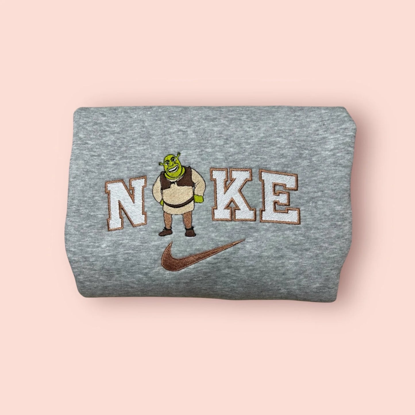 Sudadera de color gris jaspeado con un bordado de alta calidad de Shrek. El diseño presenta al ogro más famoso de DreamWorks con su clásica chaleco y sonrisa socarrona, integrado en letras estilo varsity (universitario). Una prenda imprescindible para fans de la cultura pop y los memes. Ropa personalizada divertida, cálida y cómoda. El regalo perfecto para tu novio, mejor amigo o para cualquiera que quiera defender su ciénaga con estilo.