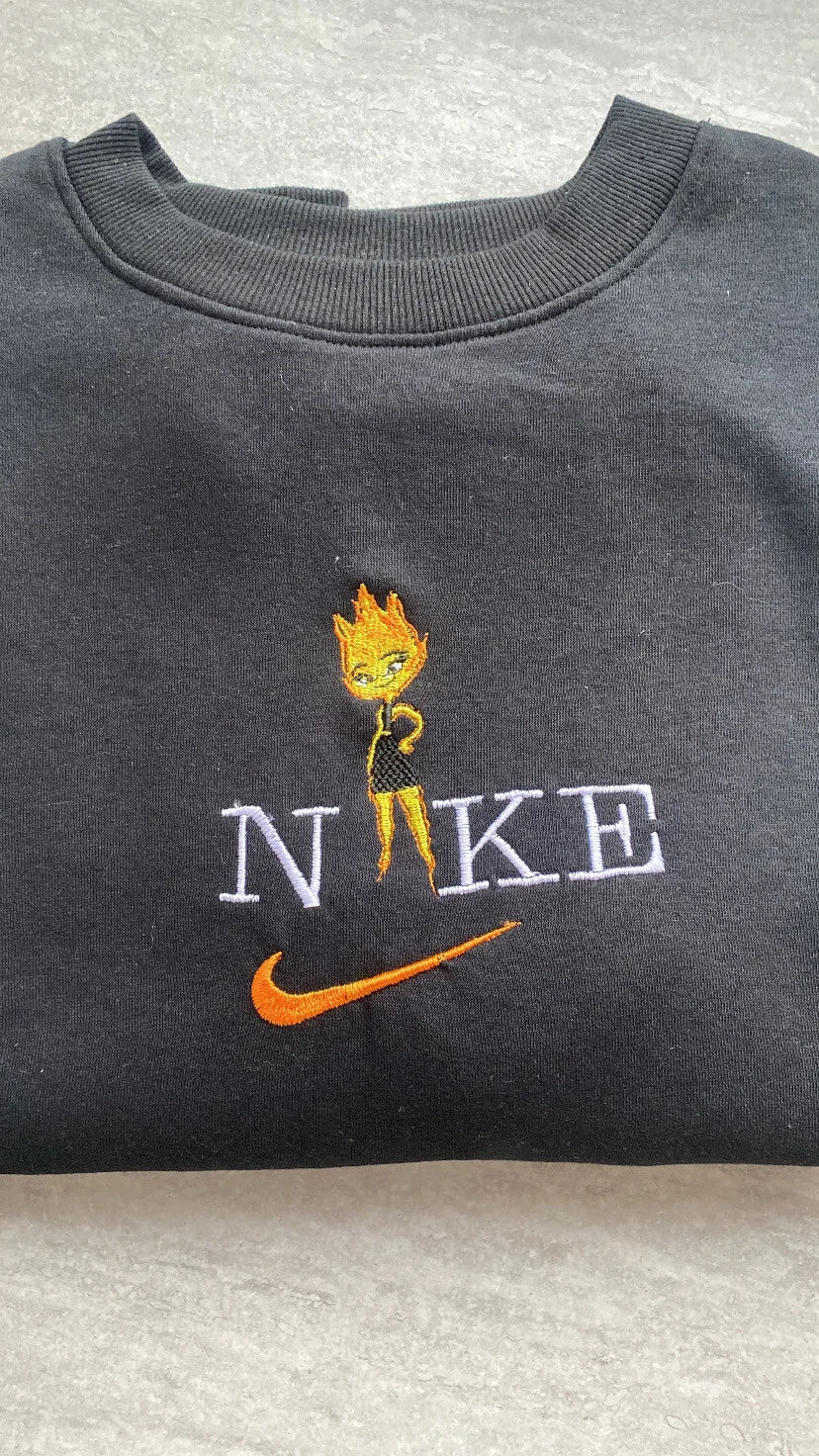 Sudadera de color negro intenso con un vibrante bordado de Candela (Ember Lumen) de la película de Pixar, Elemental. El diseño muestra al apasionado personaje de fuego con sus llamas naranjas y amarillas, integrada en letras estilo universitario. Ropa personalizada de alta calidad, perfecta para chicas con carácter. El regalo ideal para combinar con la sudadera de Nilo y demostrar que los opuestos se atraen.