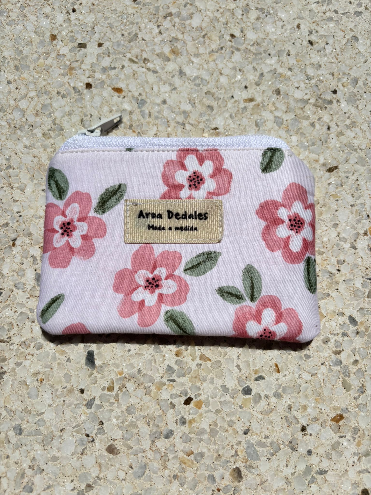Monedero blanco con estampado de flores rosas y verdes, cierre de cremallera metálica, hecho a mano por Aroa Dedales. Ideal para uso diario o como regalo artesanal.