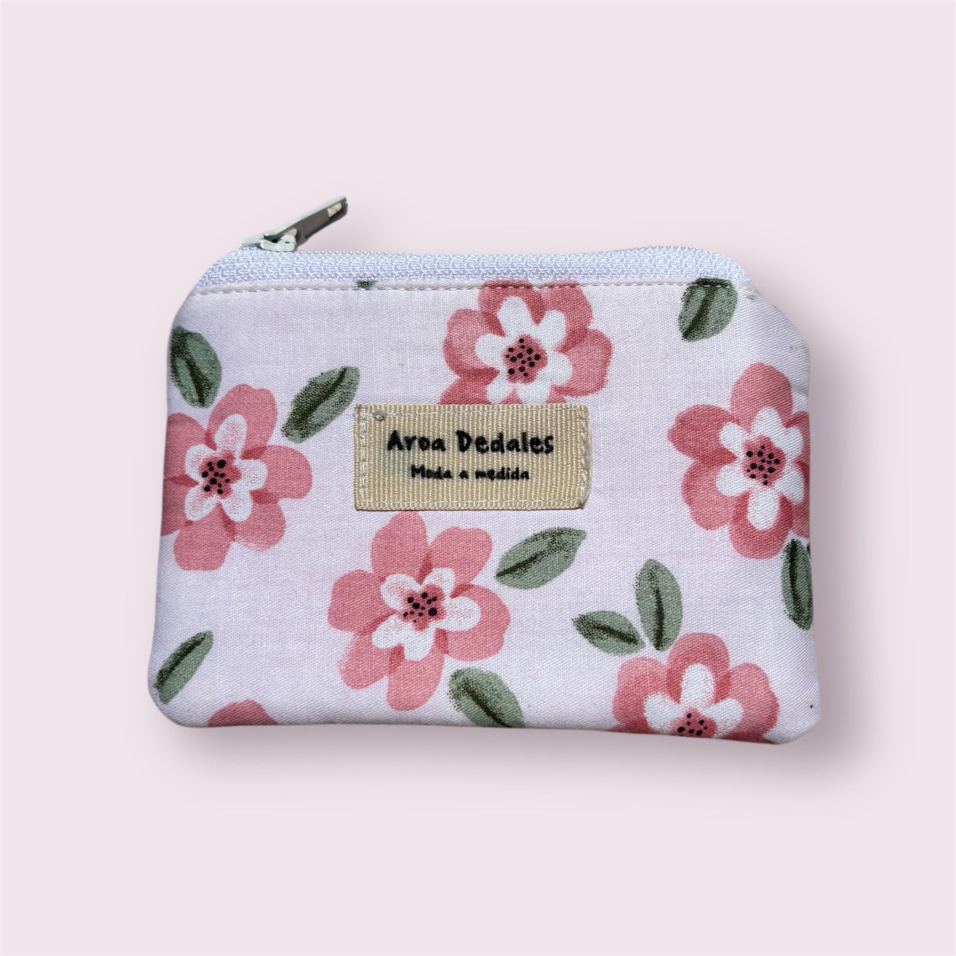 Monedero blanco con estampado de flores rosas y verdes, cierre de cremallera metálica, hecho a mano por Aroa Dedales. Ideal para uso diario o como regalo artesanal.