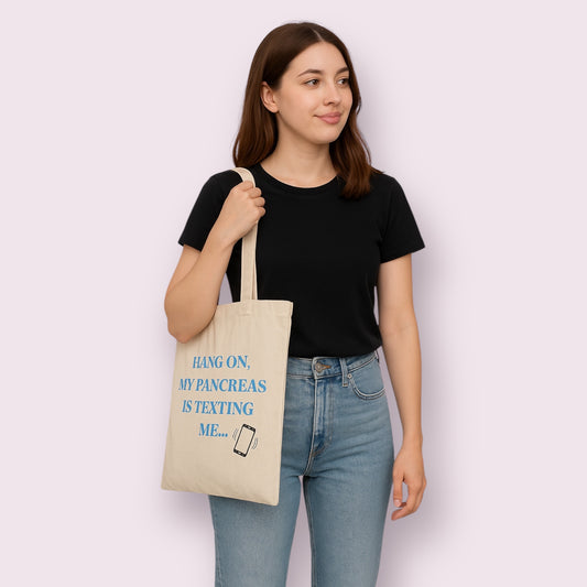 Bolso tote beige con frase divertida "Hang on, my pancreas is texting me", ideal para personas con diabetes tipo 1 que utilizan sensores o bombas de insulina y quieren mostrar su estilo con humor. Mujer joven vestida de negro lo luce con naturalidad, diseño exclusivo de Aroa Dedales