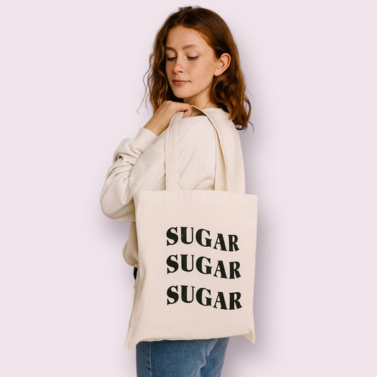 Bolso tote de algodón con el mensaje “SUGAR SUGAR SUGAR” en letras grandes negras, ideal para personas con diabetes tipo 1 que buscan un toque divertido y moderno. Mujer joven con sudadera clara y vaqueros lo lleva con estilo casual. Diseño original y con mensaje, creado por Aroa Dedales