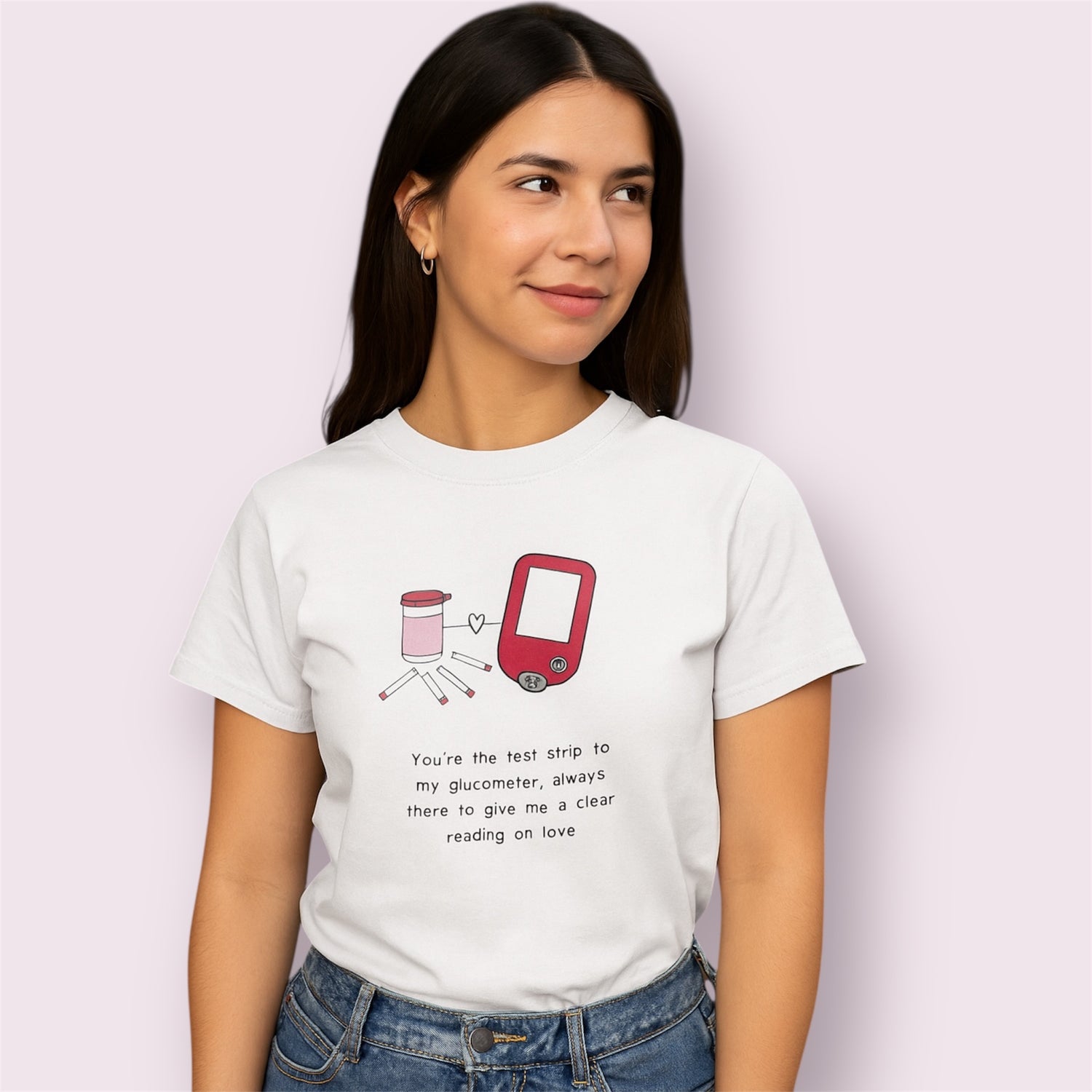 Camiseta blanca con ilustración de glucómetro y tiras reactivas junto al texto "You're the test strip to my glucometer, always there to give me a clear reading on love", modelo mujer con fondo rosa claro