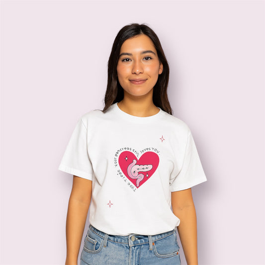 Camiseta blanca para personas con diabetes tipo 1, con diseño de páncreas dentro de un corazón rosa y texto "Your pancreas still loves you", modelo mujer sonriente