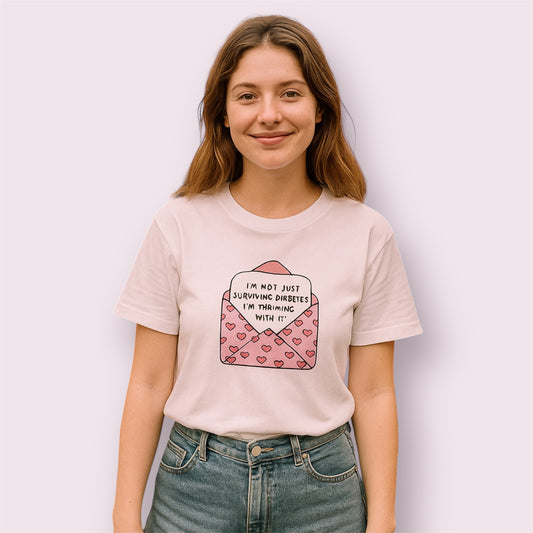 Camiseta blanca con diseño de sobre rosa y corazones con el mensaje "I'm not just surviving diabetes, I'm thriving with it", modelo mujer sonriendo, fondo rosa claro