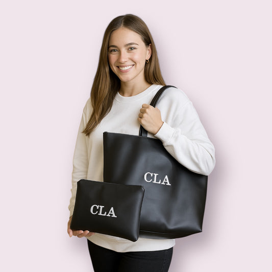 Mujer sonriente sujetando conjunto de bolso tote grande y neceser en color negro, ambos con iniciales CLA bordadas en blanco, ideal como regalo personalizado, elegante y funcional, disponible en la tienda Aroa Dedales