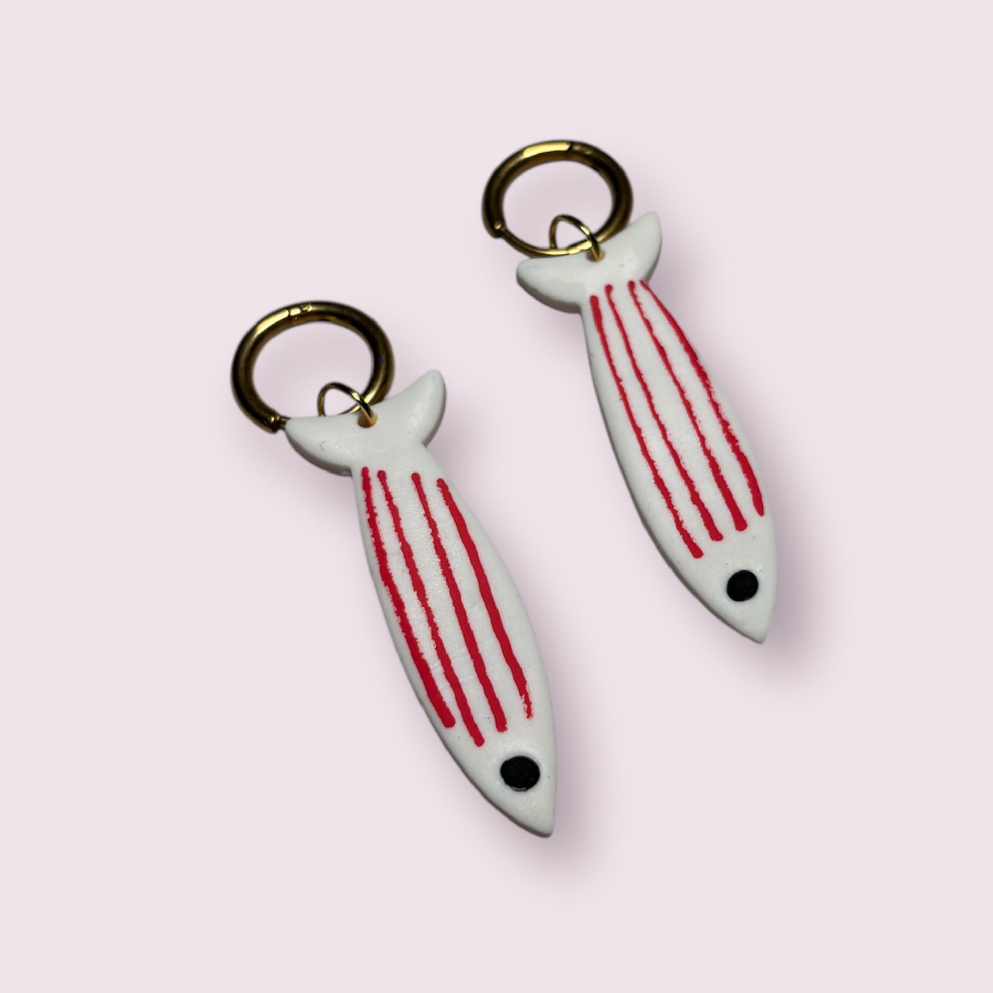 Pendientes originales con forma de sardina blanca y rayas rojas, hechos a mano con arcilla polimérica por Aroa Dedales. Diseño artesanal con aro metálico dorado, ligeros y cómodos para el día a día. Perfectos como complemento veraniego con estilo mediterráneo, ideales para amantes del mar, la moda creativa y los accesorios únicos. Regalo hecho en España para mujeres que buscan joyas diferentes y divertidas con inspiración marina.