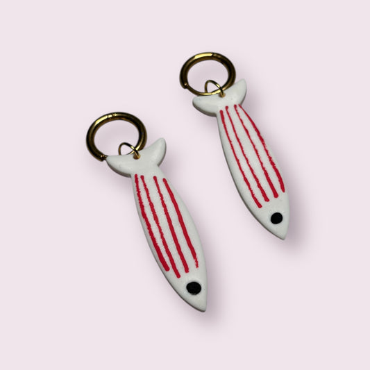 Pendientes originales con forma de sardina blanca y rayas rojas, hechos a mano con arcilla polimérica por Aroa Dedales. Diseño artesanal con aro metálico dorado, ligeros y cómodos para el día a día. Perfectos como complemento veraniego con estilo mediterráneo, ideales para amantes del mar, la moda creativa y los accesorios únicos. Regalo hecho en España para mujeres que buscan joyas diferentes y divertidas con inspiración marina.
