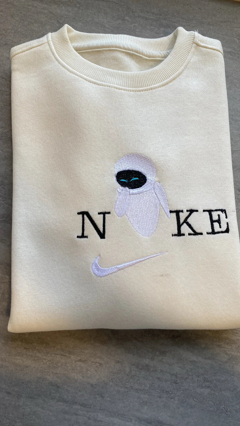 Sudadera de color beige crema con un bordado elegante y minimalista de Eva (EVE), el robot futurista de la película WALL-E. El diseño muestra a Eva riendo con sus brillantes ojos azules, integrada en un logo de estilo deportivo con letras negras. Ropa personalizada de Disney Pixar de alta calidad. El regalo imprescindible para parejas que quieren hacer "matching outfits" combinándola con la sudadera de WALL-E. Estilo tierno y moderno.