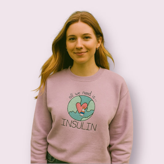 Mujer joven con sudadera lila con diseño bordado de un corazón y un vial de insulina sobre un mapa del mundo con la frase "All we need is insulin", prenda original para personas con diabetes tipo 1, moda con mensaje positivo y visibilidad para la diabetes