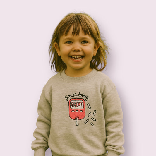 Niña sonriente con sudadera infantil gris claro con diseño de glucómetro rosa y mensaje motivador "You're doing great", ropa para niños con diabetes tipo 1, ideal para visibilizar la diabetes con estilo y ternura