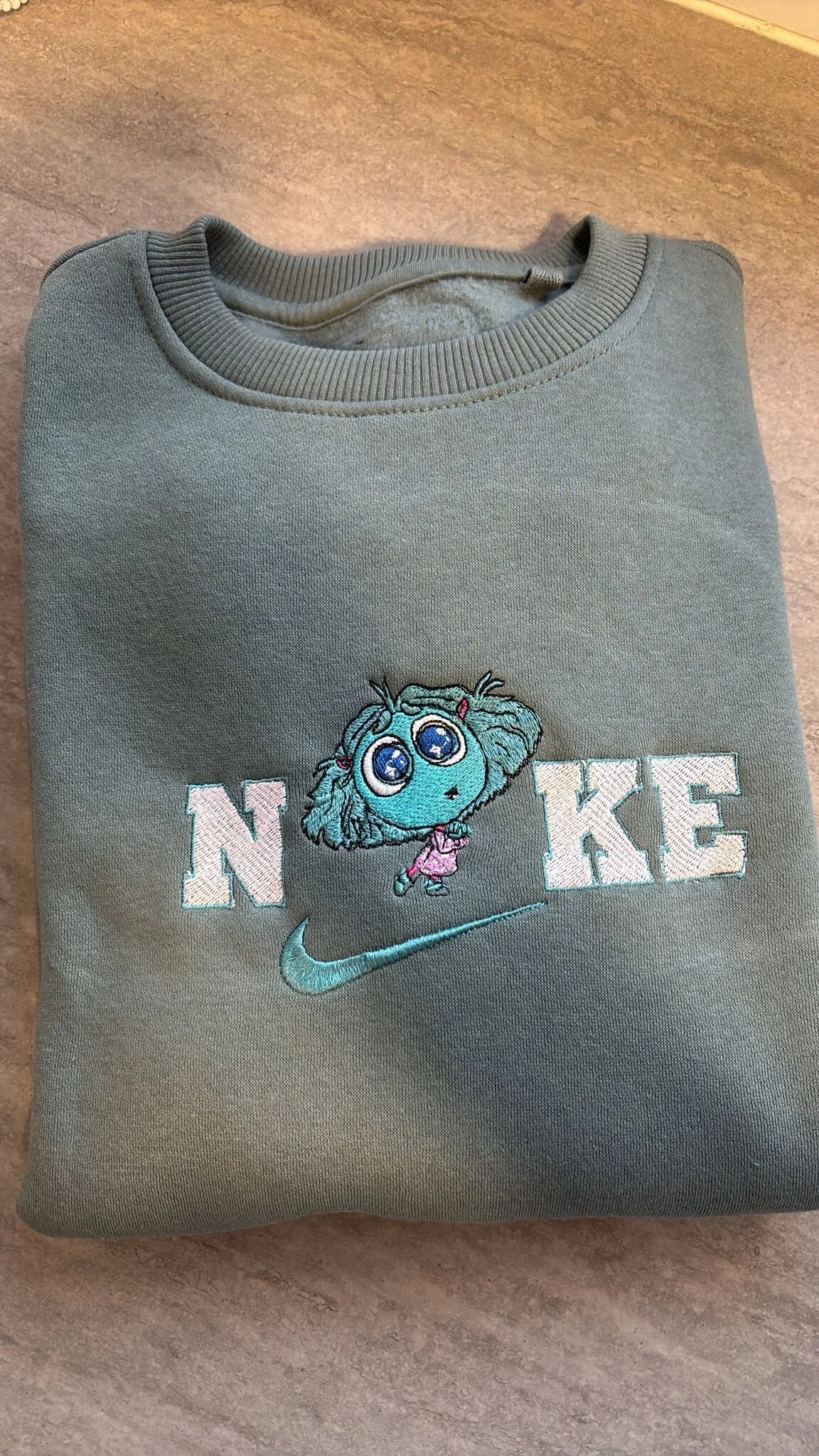 Sudadera color verde agua oscuro con bordado super tierno de Envidia (Envy), el nuevo personaje de Inside Out 2. Diseño con ojos grandes brillantes y detalles en hilo turquesa y rosa. Ropa personalizada de la película Del Revés 2. Una prenda dulce y cómoda, disponible para comprar online. Perfecta para amantes de los diseños kawaii y Disney.