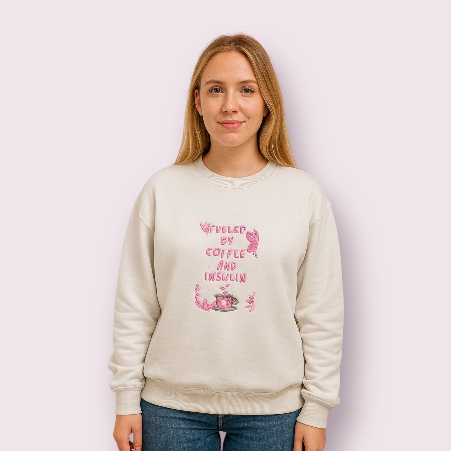 Mujer con sudadera beige con diseño bordado en hilo rosa con la frase "Fueled by coffee and insulin", pensada para personas con diabetes tipo 1 que aman el café, combinando estilo, salud y reivindicación personal con un toque tierno y reivindicativo, ideal para destacar en redes sociales y concienciar sobre la vida con diabetes