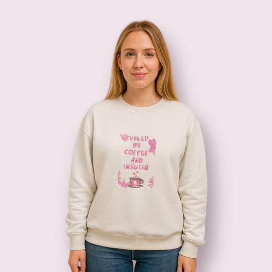 Mujer con sudadera beige con diseño bordado en hilo rosa con la frase "Fueled by coffee and insulin", pensada para personas con diabetes tipo 1 que aman el café, combinando estilo, salud y reivindicación personal con un toque tierno y reivindicativo, ideal para destacar en redes sociales y concienciar sobre la vida con diabetes