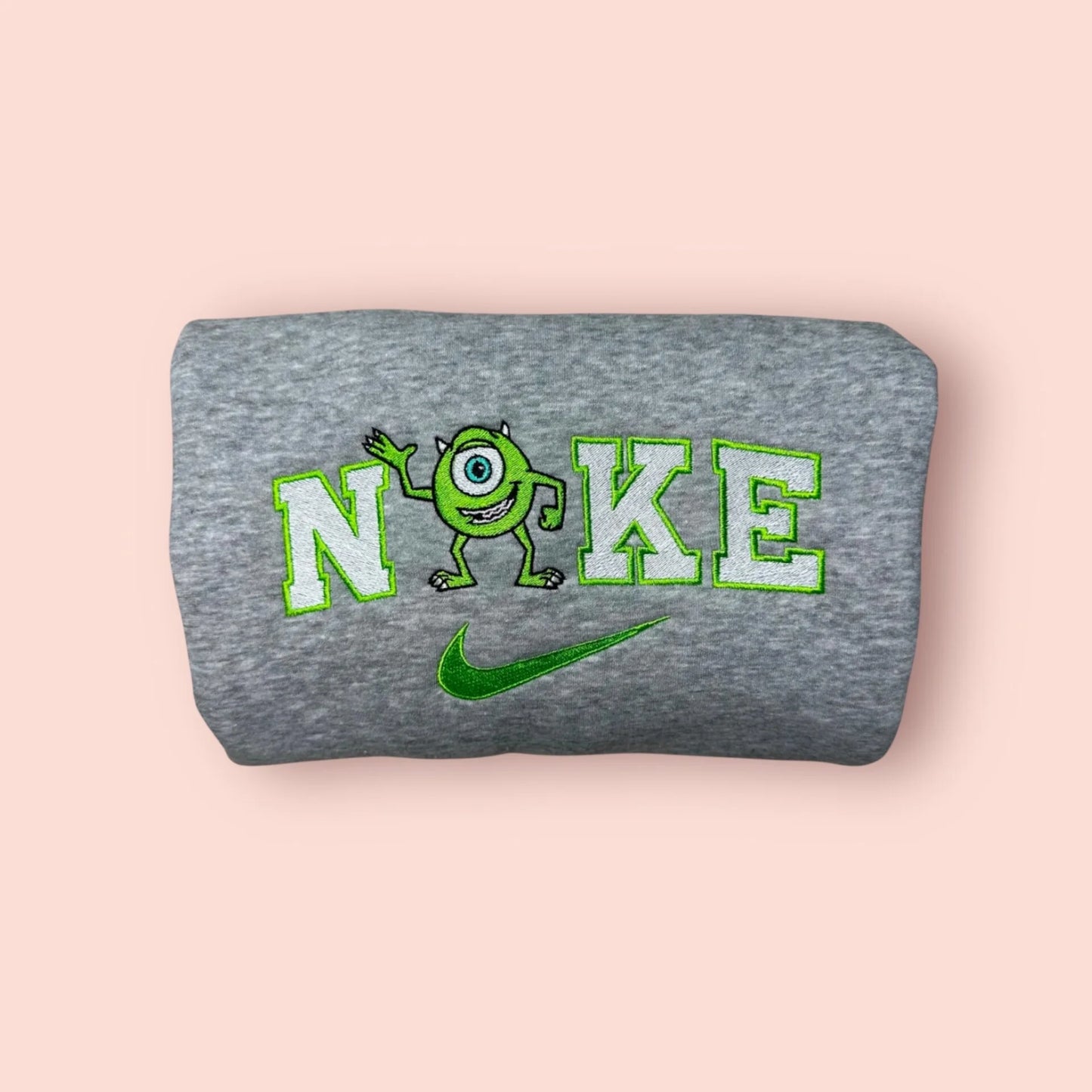 Sudadera de color gris jaspeado con un bordado verde neón de Mike Wazowski. El diseño muestra al divertido monstruo de un ojo saludando, integrado en letras estilo universitario. Ropa personalizada de Monstruos S.A. (Monsters, Inc.), perfecta para regalar a tu mejor amigo. Combínala con la sudadera de Sulley para crear el "matching outfit" definitivo del mejor equipo de sustos.