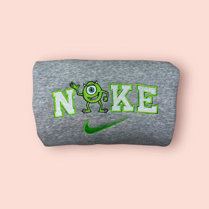 Sudadera de color gris jaspeado con un bordado verde neón de Mike Wazowski. El diseño muestra al divertido monstruo de un ojo saludando, integrado en letras estilo universitario. Ropa personalizada de Monstruos S.A. (Monsters, Inc.), perfecta para regalar a tu mejor amigo. Combínala con la sudadera de Sulley para crear el "matching outfit" definitivo del mejor equipo de sustos.