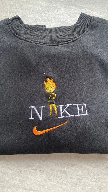 Sudadera de color negro intenso con un vibrante bordado de Candela (Ember Lumen) de la película de Pixar, Elemental. El diseño muestra al apasionado personaje de fuego con sus llamas naranjas y amarillas, integrada en letras estilo universitario. Ropa personalizada de alta calidad, perfecta para chicas con carácter. El regalo ideal para combinar con la sudadera de Nilo y demostrar que los opuestos se atraen.
