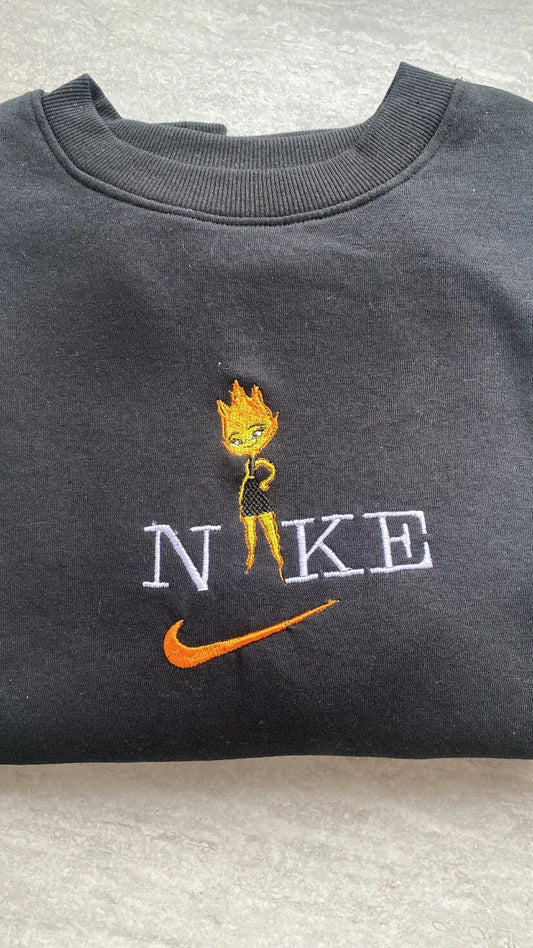 Sudadera de color negro intenso con un vibrante bordado de Candela (Ember Lumen) de la película de Pixar, Elemental. El diseño muestra al apasionado personaje de fuego con sus llamas naranjas y amarillas, integrada en letras estilo universitario. Ropa personalizada de alta calidad, perfecta para chicas con carácter. El regalo ideal para combinar con la sudadera de Nilo y demostrar que los opuestos se atraen.