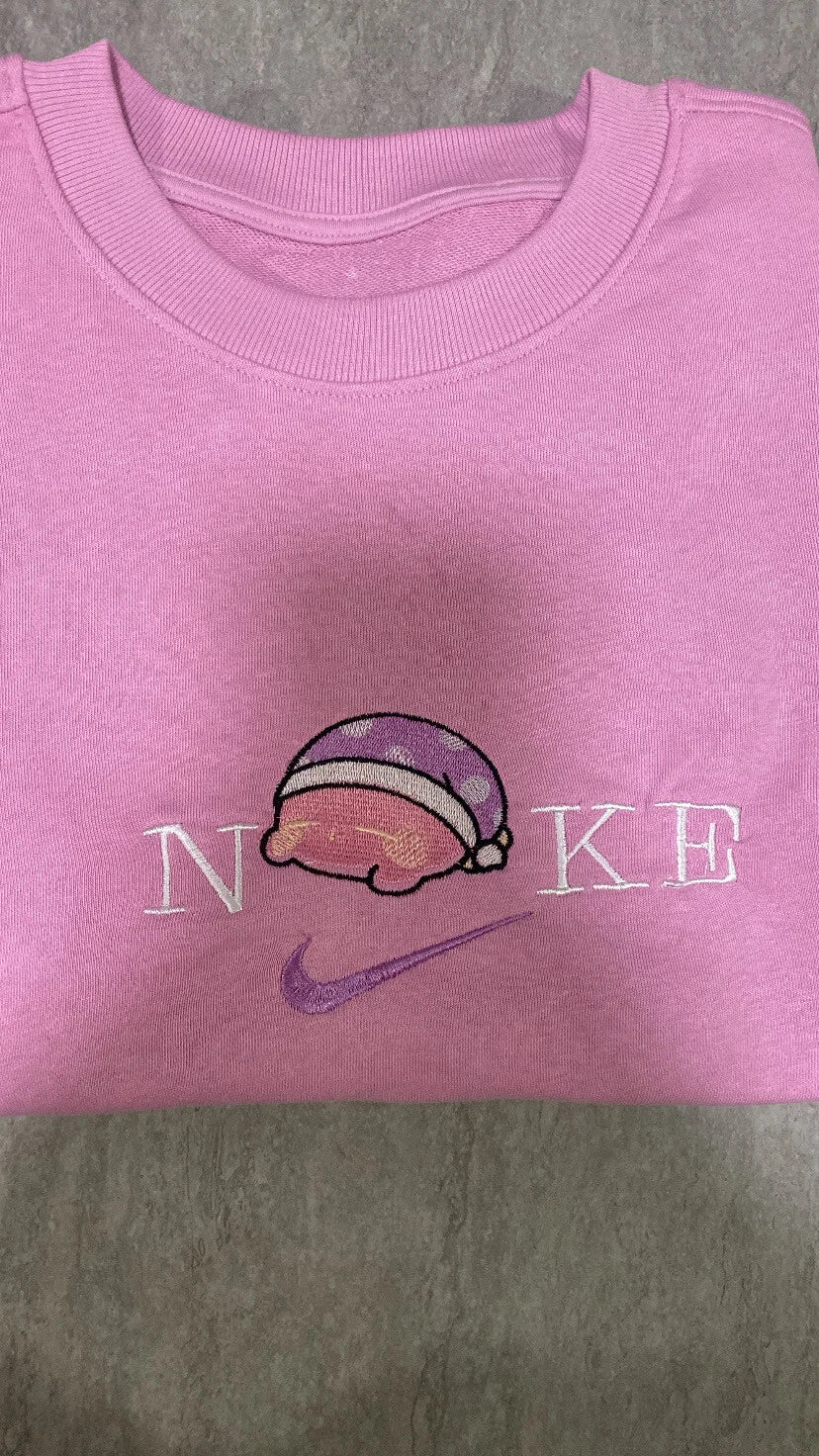 Sudadera de color rosa chicle con un bordado adorable de Kirby durmiendo la siesta. El diseño muestra al personaje de Nintendo con su icónico gorro de dormir de lunares, integrado sobre el logo con letras blancas. Ropa personalizada estilo "soft girl" o "kawaii", perfecta para outfits cómodos, tardes de videojuegos o como regalo para esa persona que ama dormir tanto como Kirby.