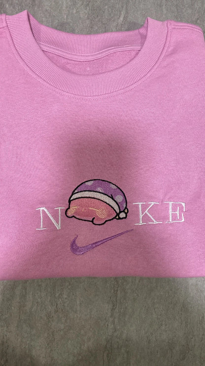 Sudadera de color rosa chicle con un bordado adorable de Kirby durmiendo la siesta. El diseño muestra al personaje de Nintendo con su icónico gorro de dormir de lunares, integrado sobre el logo con letras blancas. Ropa personalizada estilo "soft girl" o "kawaii", perfecta para outfits cómodos, tardes de videojuegos o como regalo para esa persona que ama dormir tanto como Kirby.