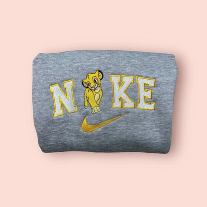 Sudadera de color gris jaspeado con un bordado dulce y detallado de Nala (cachorra) de El Rey León. Diseño con hilos en tonos crema y dorados sobre letras estilo universitario. Ropa personalizada de alta calidad, suave y cálida. El regalo perfecto para ella, ideal para crear un conjunto a juego con la sudadera de Simba. Moda Disney discreta y elegante.