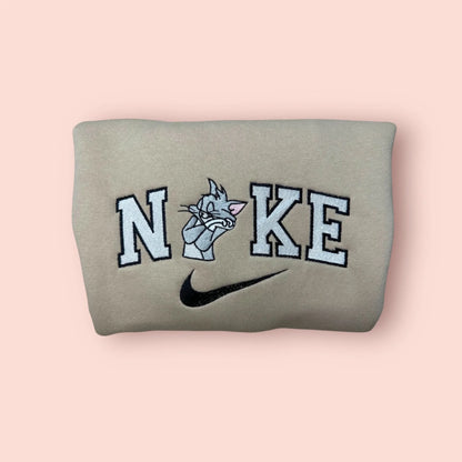 Sudadera beige con un bordado detallado del gato Tom. El personaje clásico de los dibujos animados aparece con su expresión de estar tramando algo, integrado en letras estilo varsity negras y blancas. Prenda de estilo vintage, ideal para regalar a ese amigo o pareja que siempre está metido en líos. La pieza clave para completar el dúo con la sudadera de Jerry.