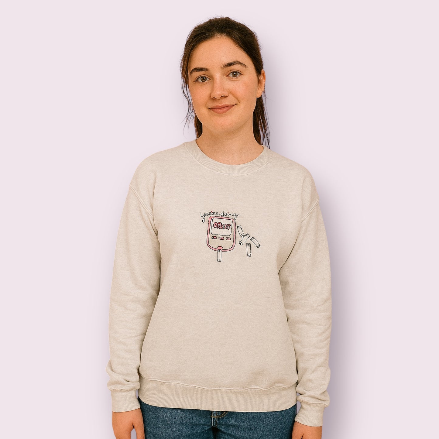 Mujer joven luciendo una sudadera beige con diseño bordado de un glucómetro rosa y la frase “You’re doing GREAT”, rodeado de tiras reactivas, pensada para visibilizar la diabetes con un mensaje positivo y motivador. Ideal para personas con diabetes tipo 1 o tipo 2 que buscan moda inclusiva, cómoda y con estilo propio