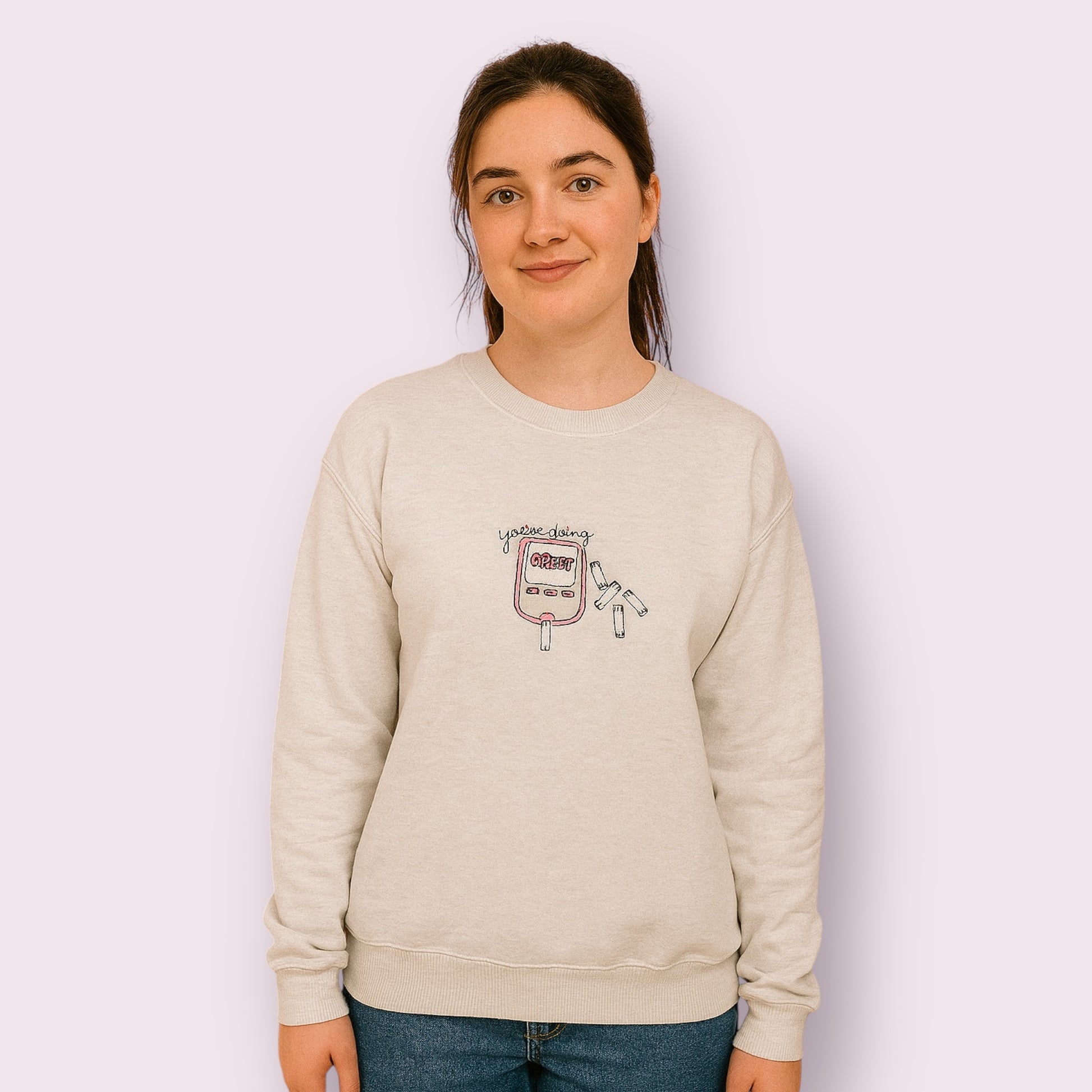 Mujer joven luciendo una sudadera beige con diseño bordado de un glucómetro rosa y la frase “You’re doing GREAT”, rodeado de tiras reactivas, pensada para visibilizar la diabetes con un mensaje positivo y motivador. Ideal para personas con diabetes tipo 1 o tipo 2 que buscan moda inclusiva, cómoda y con estilo propio