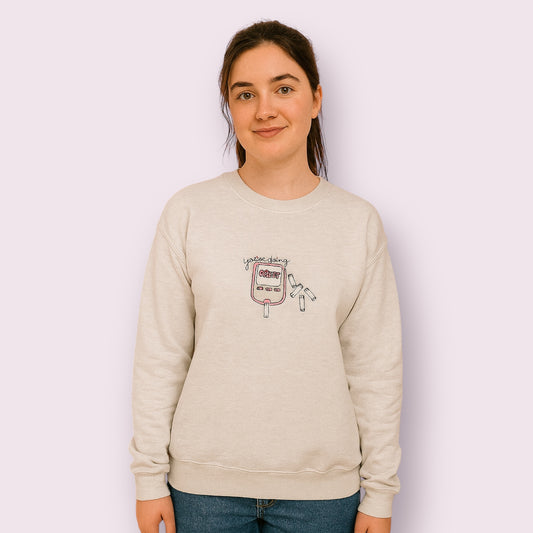 Mujer joven luciendo una sudadera beige con diseño bordado de un glucómetro rosa y la frase “You’re doing GREAT”, rodeado de tiras reactivas, pensada para visibilizar la diabetes con un mensaje positivo y motivador. Ideal para personas con diabetes tipo 1 o tipo 2 que buscan moda inclusiva, cómoda y con estilo propio