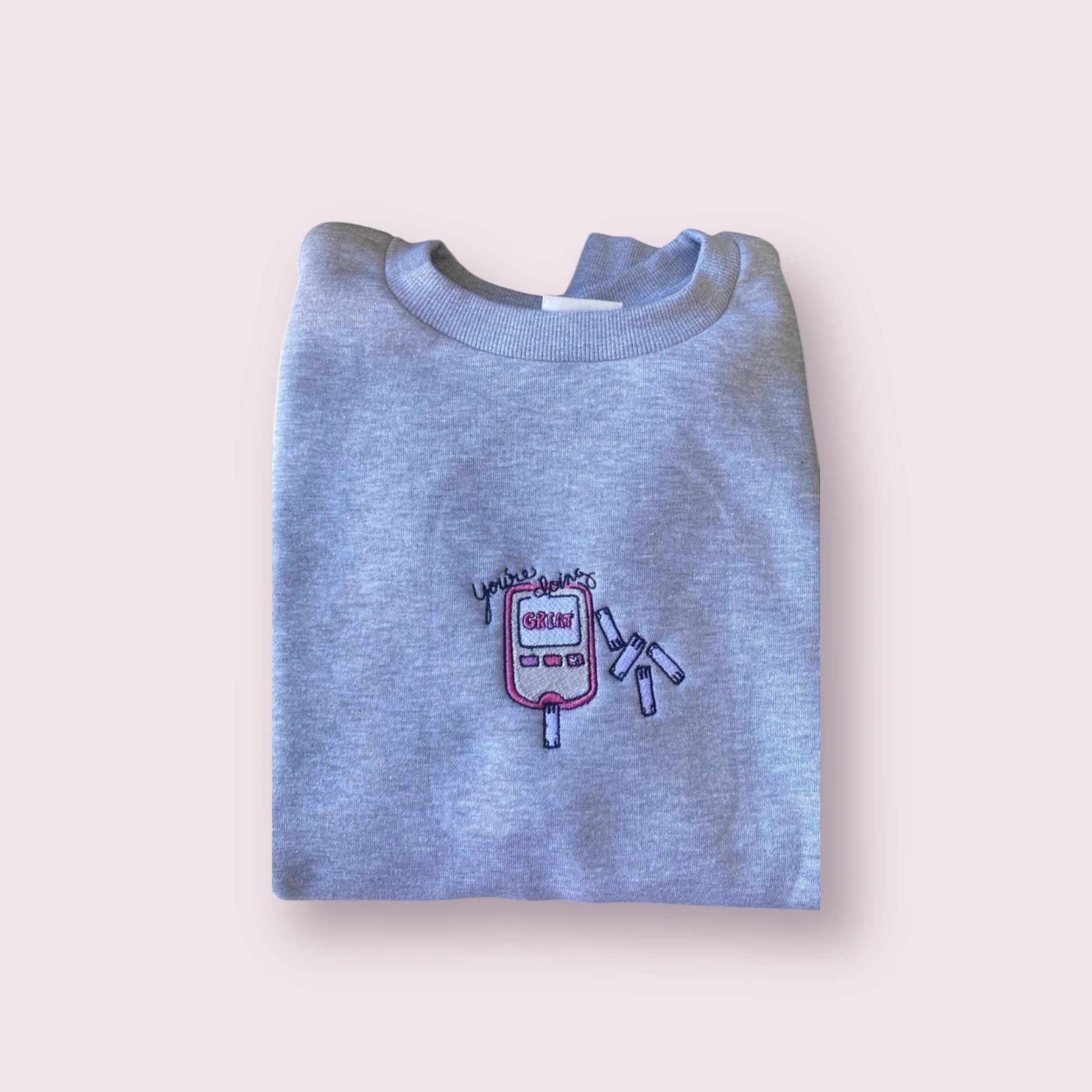 Sudadera gris con diseño bordado de un glucómetro rosa con la frase motivadora “You’re doing GREAT” y tiras reactivas alrededor, pensada para personas con diabetes tipo 1 o tipo 2 que buscan visibilizar su condición con orgullo y actitud positiva. Ideal para regalar o para vestir con humor y ternura mientras se normaliza el monitoreo diario de glucosa