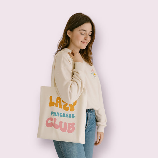 Chica joven con sudadera clara y jeans sosteniendo una bolsa tote bag de algodón color crudo con el mensaje "Lazy Pancreas Club" en colores naranja, azul y rosa. Diseño alegre para personas con diabetes tipo 1 que quieren visibilizar su condición con humor y estilo. Bolsa de tela reutilizable ideal para el día a día