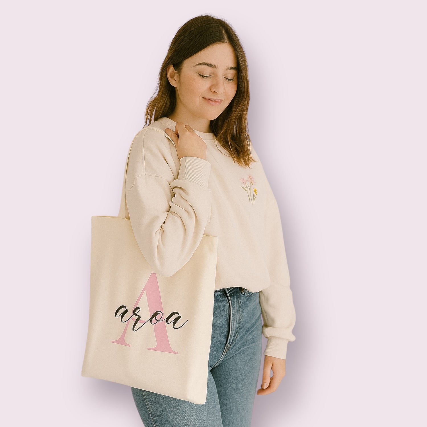 Bolsa de tela personalizada estilo tote bag con la palabra "Aroa" impresa sobre una gran letra "A" rosa pastel, sostenida por una chica joven con sudadera beige y vaqueros. Ideal como regalo original para mujeres, estudiantes o profesoras. Diseño elegante, moderno y funcional, perfecta para personas con nombre que empieza por A, amante de los productos personalizados