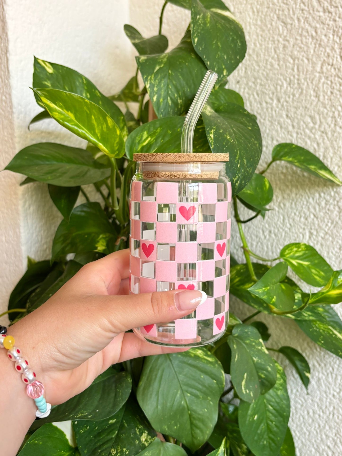 Vaso de cristal reutilizable con diseño de cuadros rosas y pequeños corazones rojos, acompañado de tapa de bambú natural y pajita de cristal. Ideal para regalar en cumpleaños, profes, amigas o enamorados. Perfecto para café helado, smoothies o bebidas frías. Un complemento bonito, ecológico y práctico que destaca por su estilo dulce y romántico. Artesanal, funcional y sostenible.